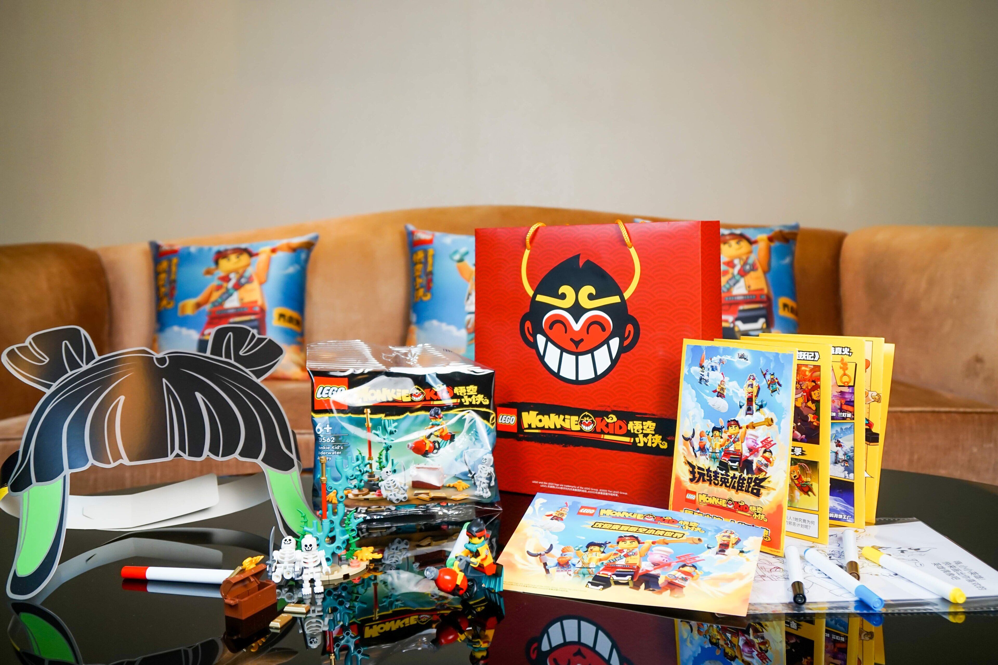Una mesa con varios juegos y accesorios LEGO, una bolsa roja con un diseño de rostro, y un sofá con almohadas al fondo.