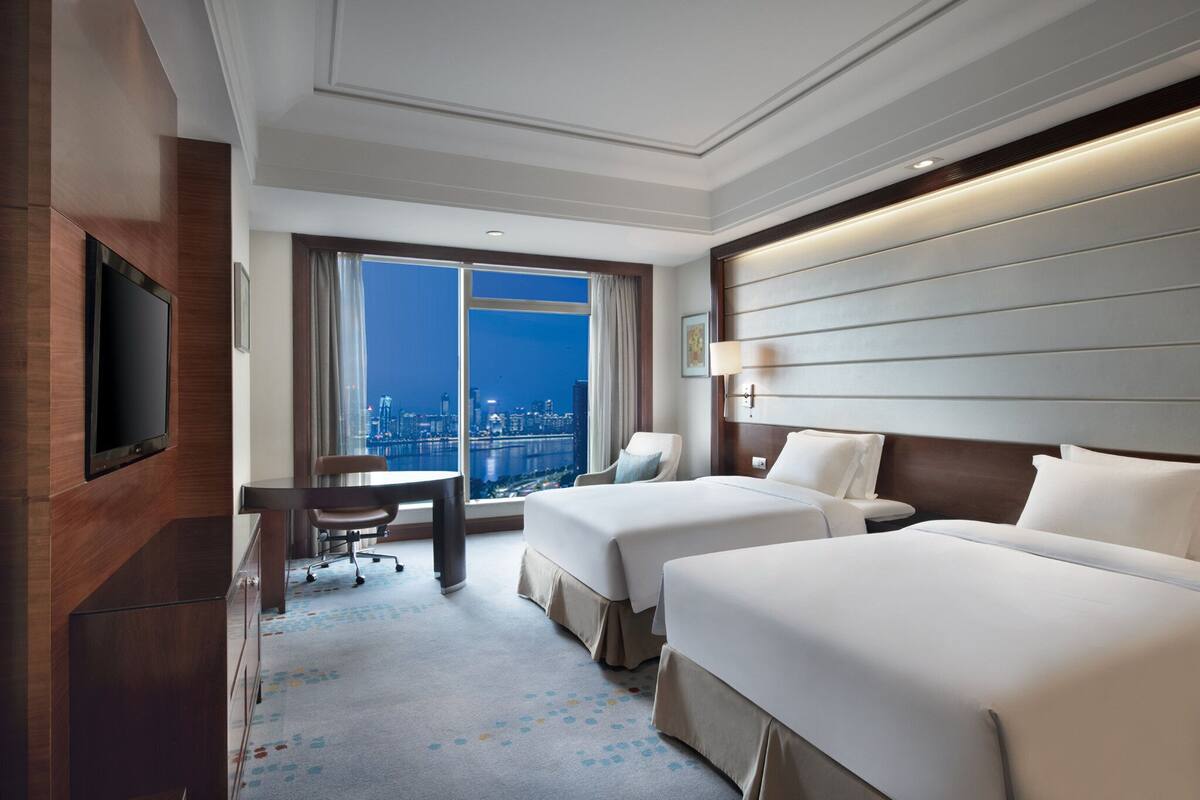 A hotel room with two beds, a desk, a chair, a TV, and a large window with a city view.