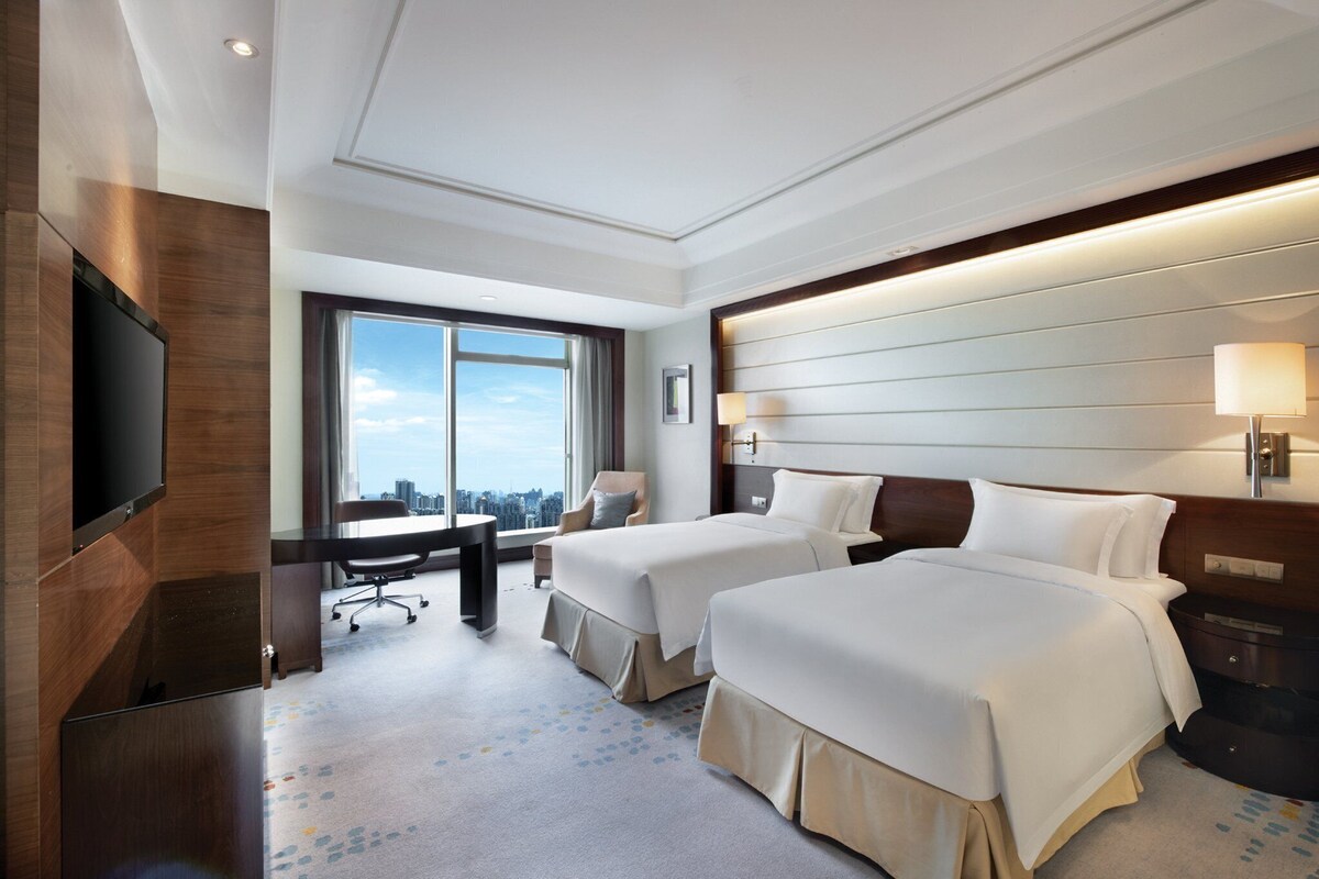 A hotel room with two beds, a desk, a chair, a TV, and a large window offering a city view.