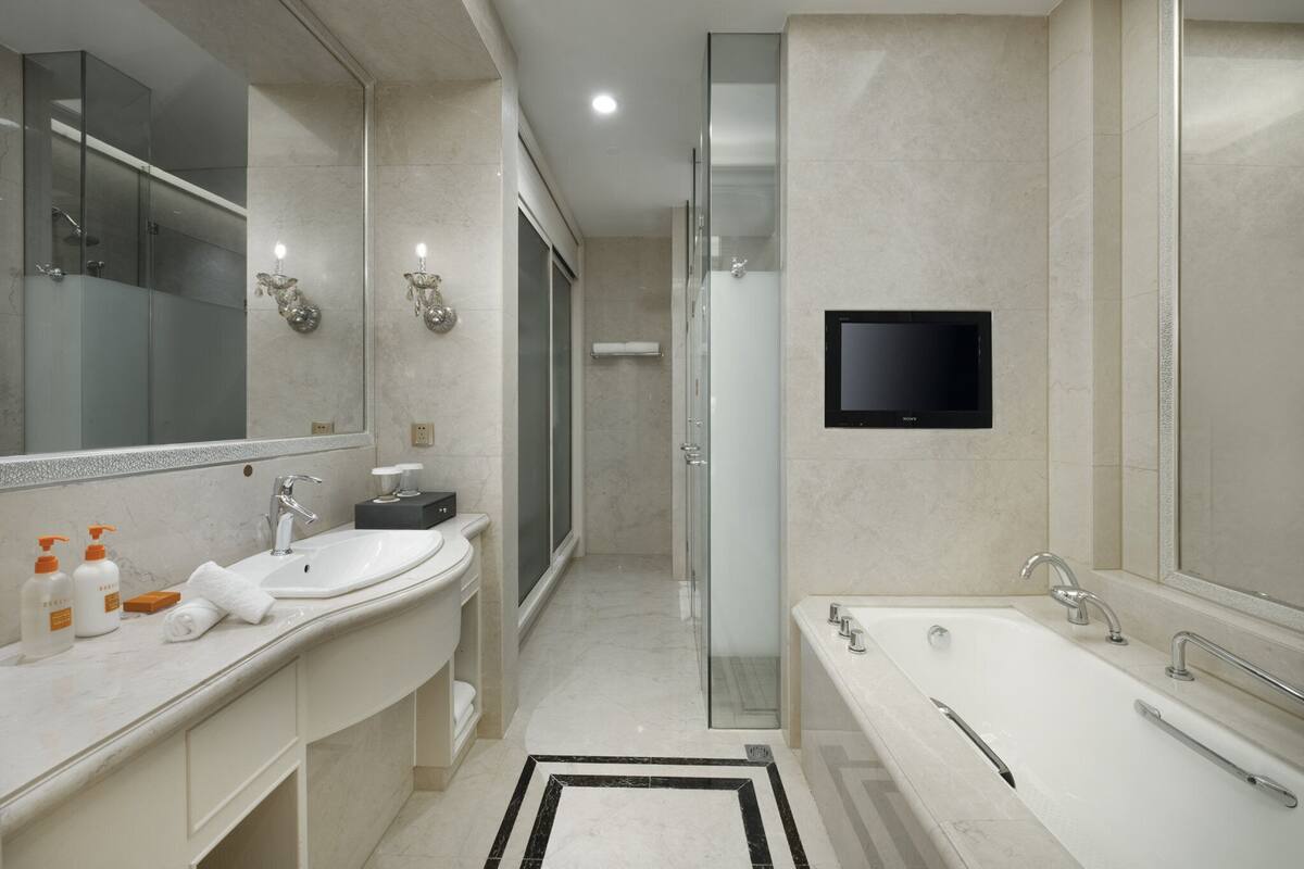 A modern bathroom with a large mirror, two sinks, a bathtub, and a TV mounted on the wall.