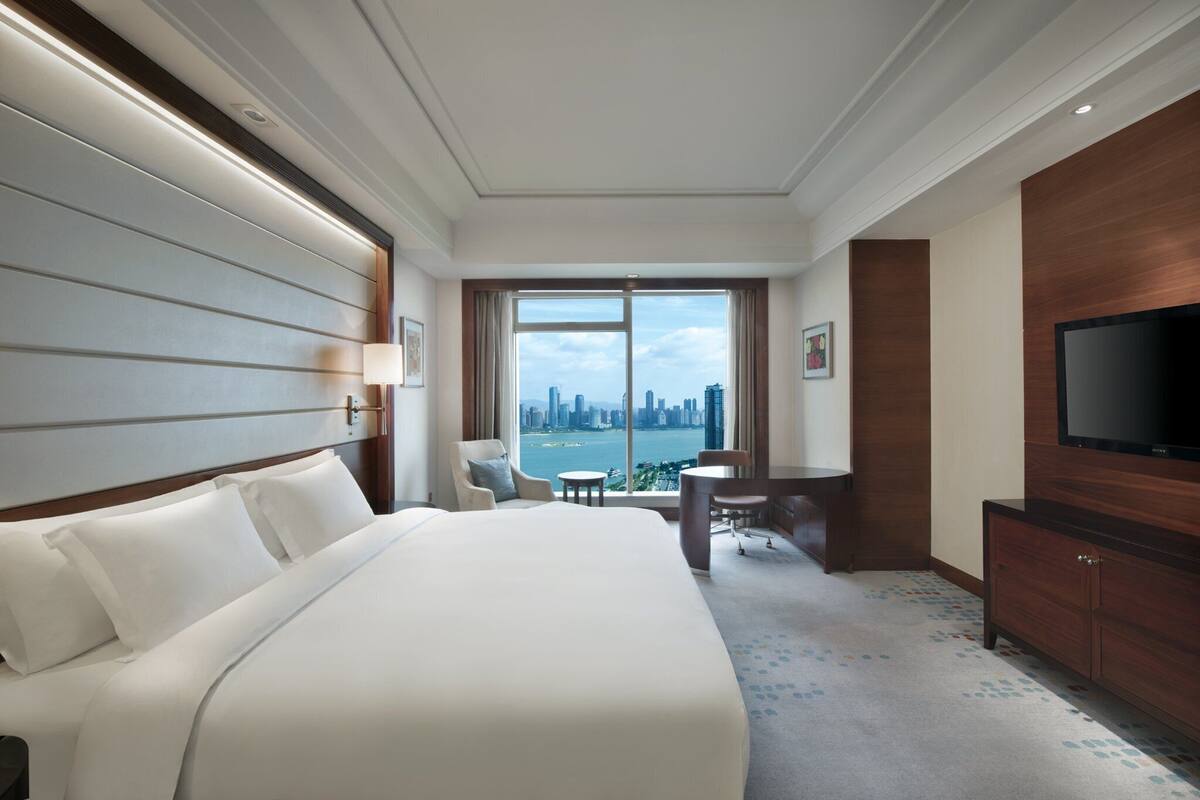 A hotel room with a large bed, a desk, a chair, a TV, and a view of a city skyline.