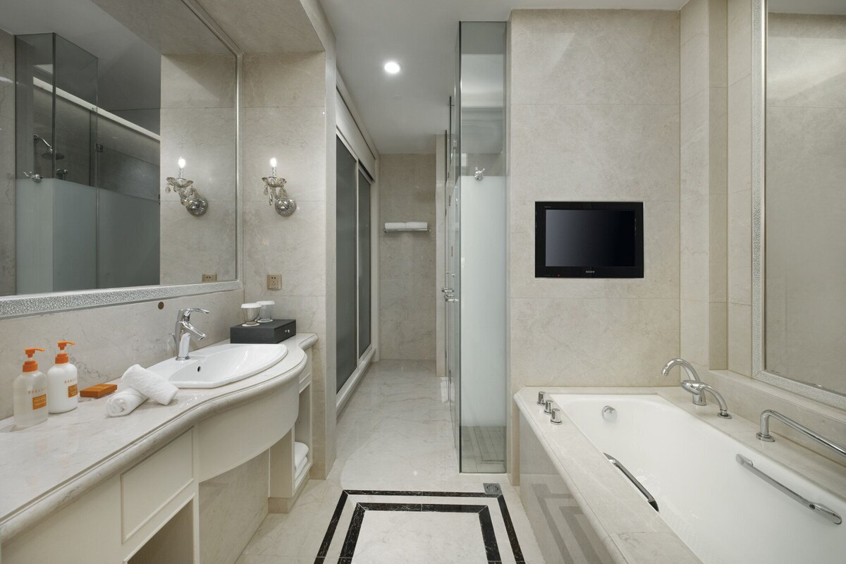 A modern bathroom with a large mirror, two sinks, a bathtub, and a TV mounted on the wall.