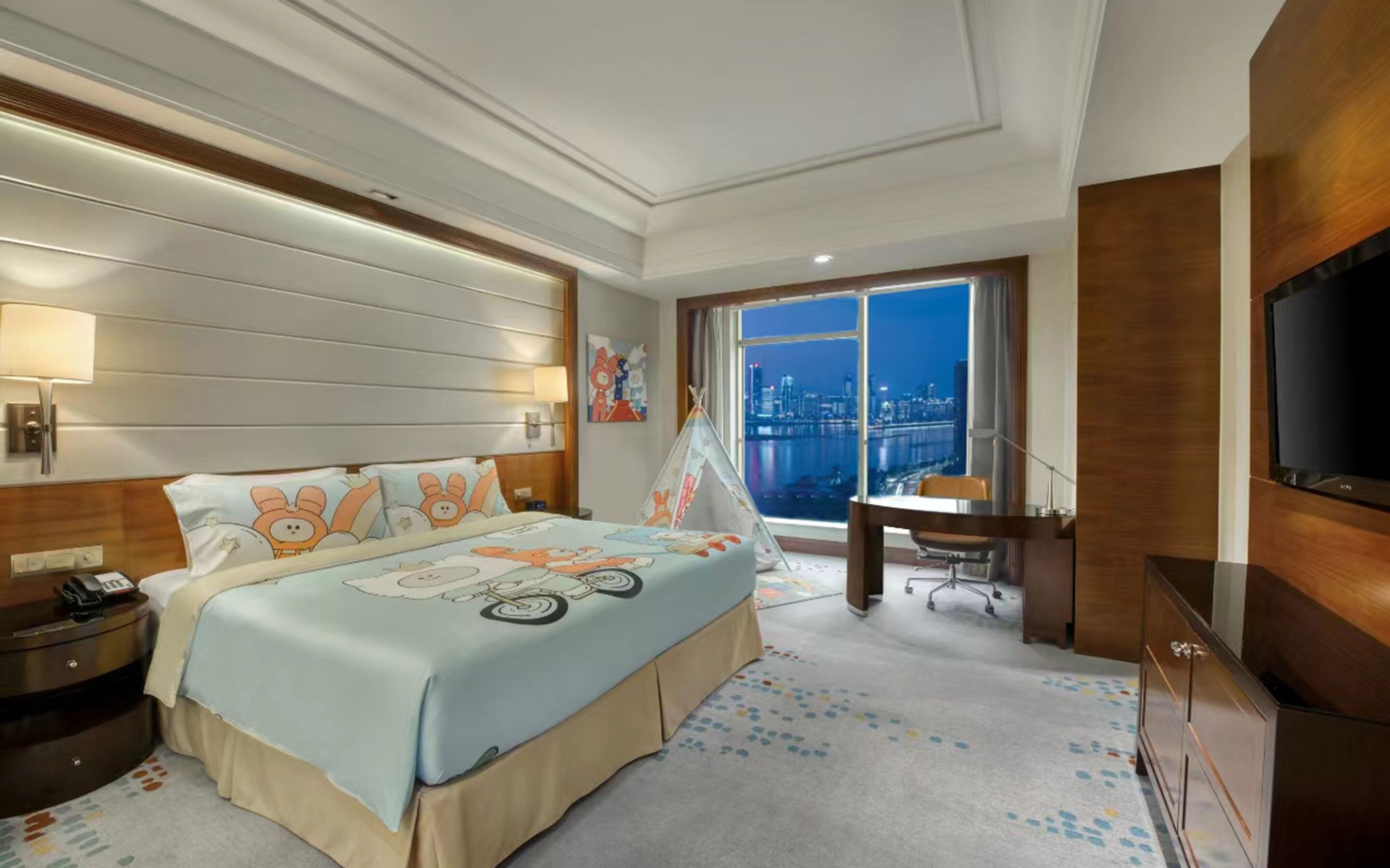 Habitación de hotel con una cama decorada con personajes de caricatura, vistas de la ciudad a través de ventanas grandes, un escritorio con una silla y un televisor montado en la pared.