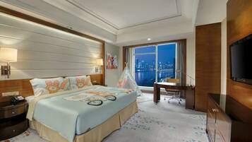Habitación de hotel con una cama decorada con personajes de caricatura, vistas de la ciudad a través de ventanas grandes, un escritorio con una silla y un televisor montado en la pared.