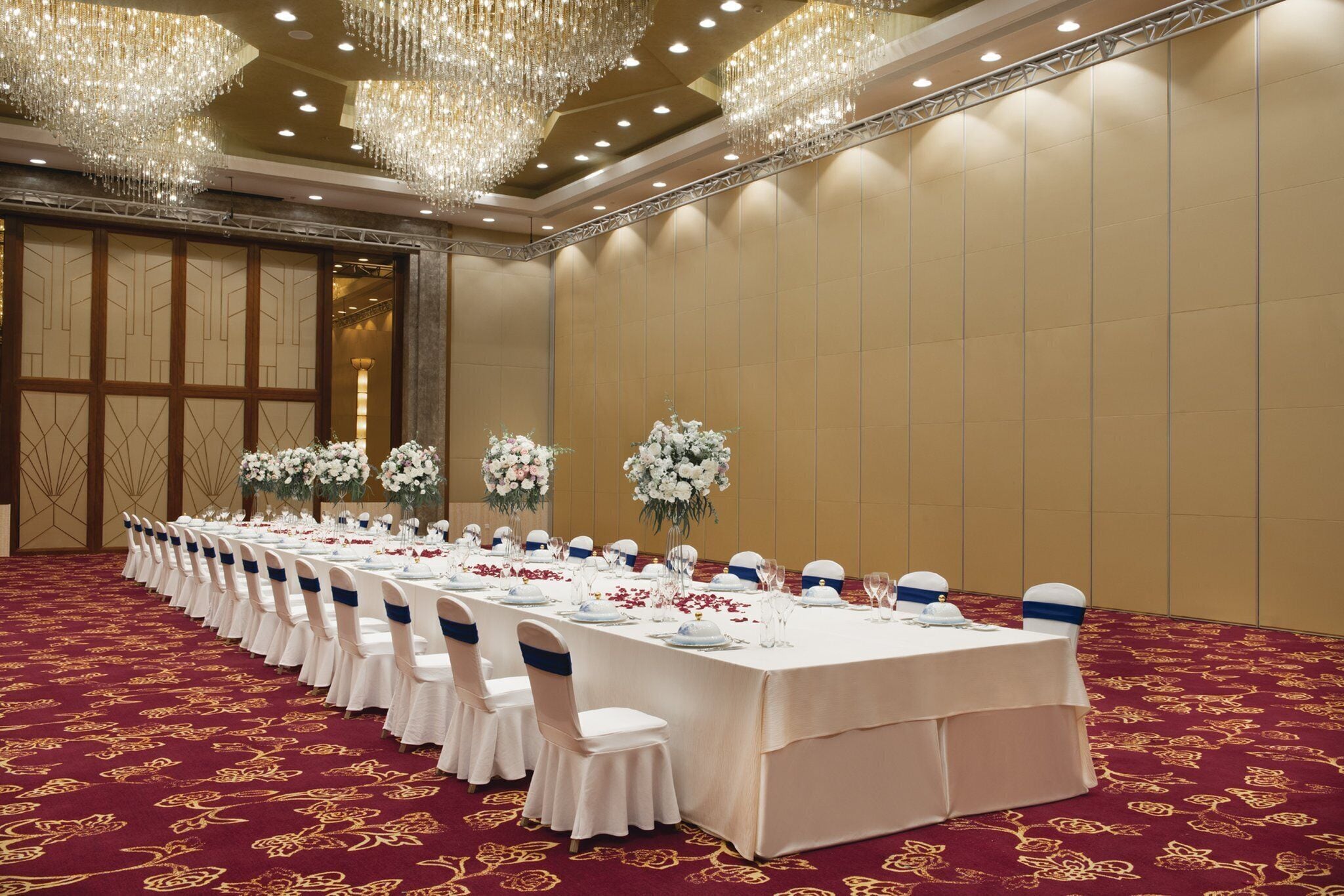 Una sala de banquet amb una taula llarga preparada per a un esdeveniment formal, flanquejada per alts arranjaments florals i un ostentós candelabrum.