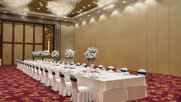 Una sala de banquet amb una taula llarga preparada per a un esdeveniment formal, flanquejada per alts arranjaments florals i un ostentós candelabrum.