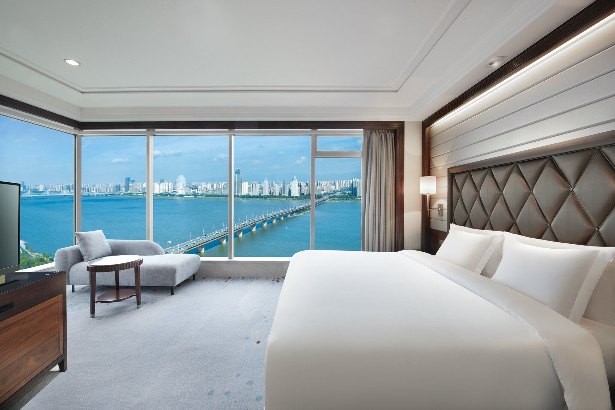 A hotel room with a large bed, a sofa, a TV, and a view of a city skyline and bridge.