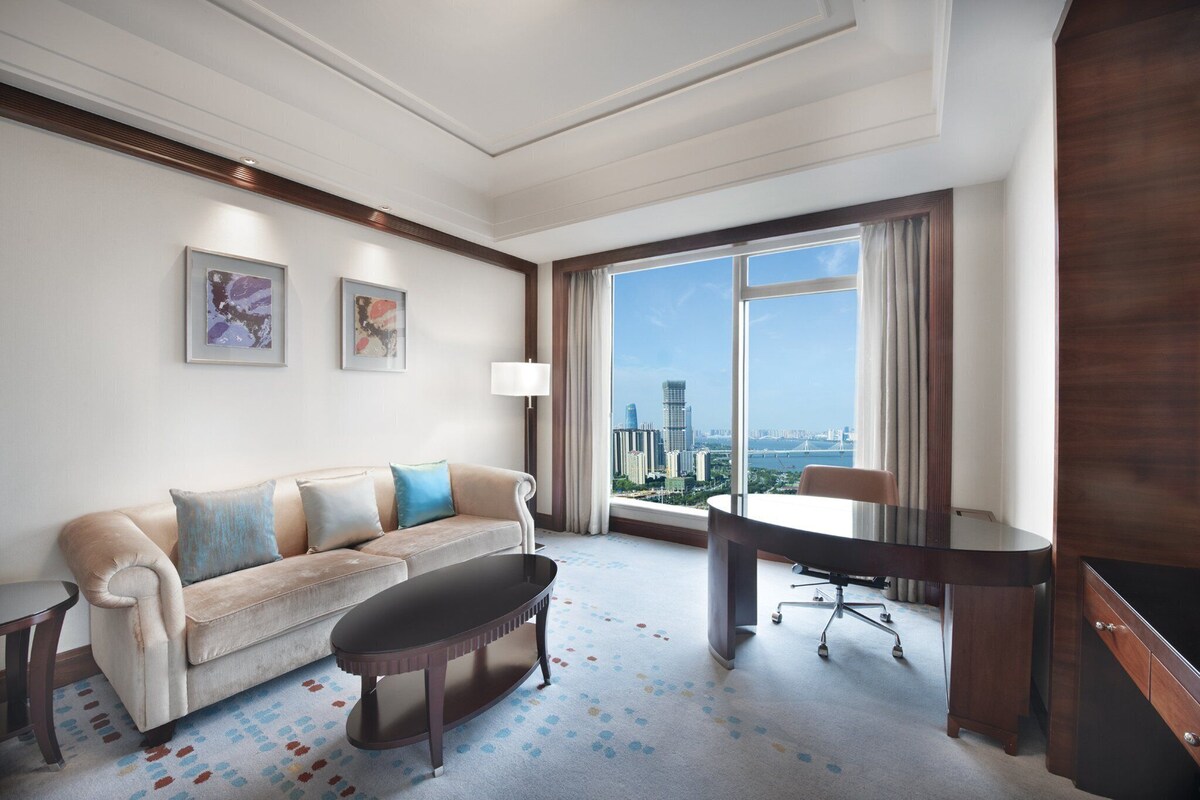 A hotel room with a large window, a sofa, a desk, and a TV.