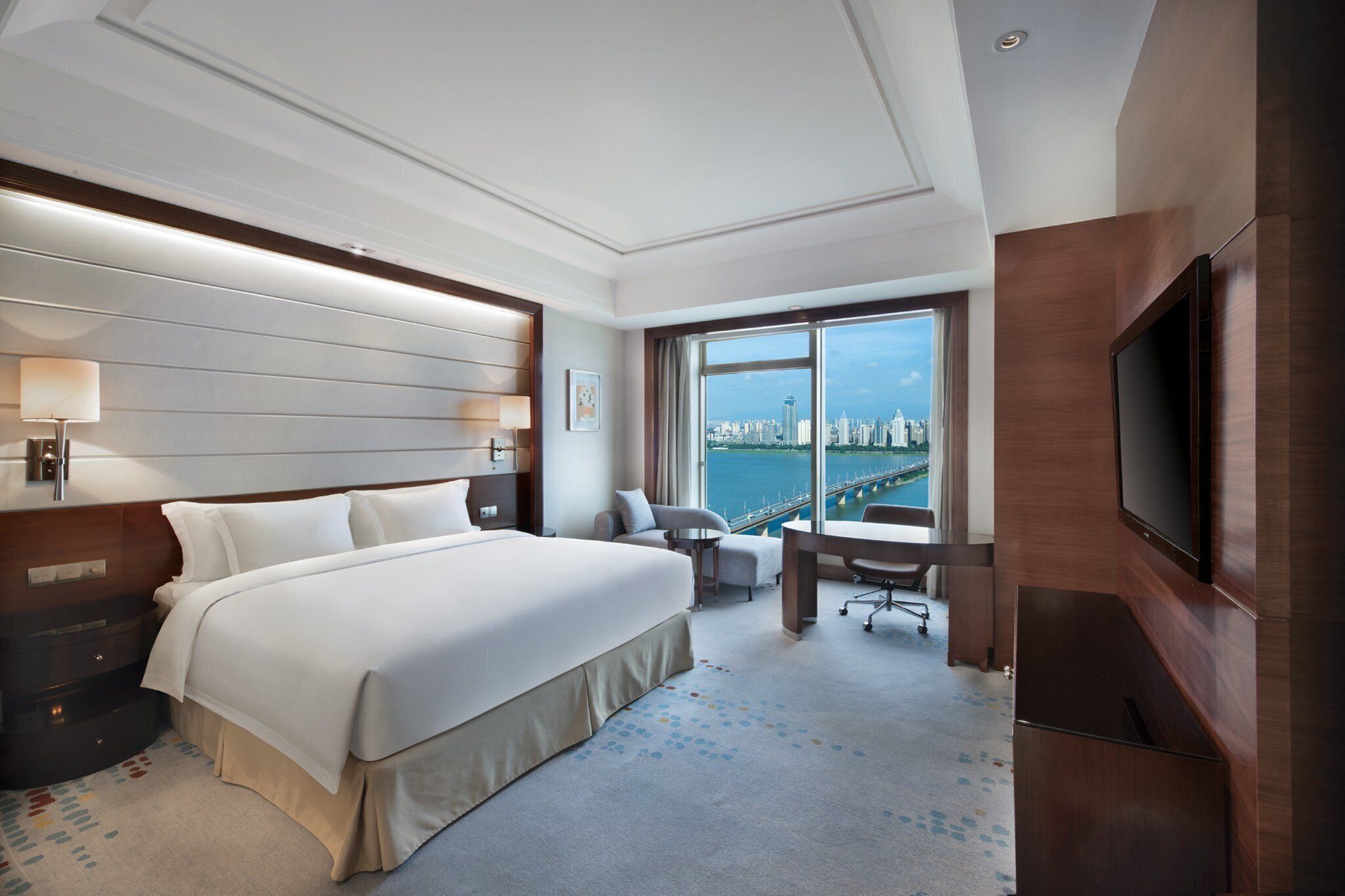 A hotel room with a large bed, a desk, a chair, a TV, and a view of a cityscape.