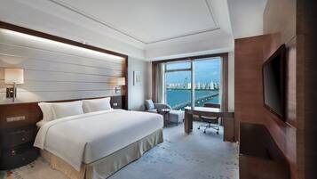 A hotel room with a large bed, a desk, a chair, a TV, and a view of a cityscape.