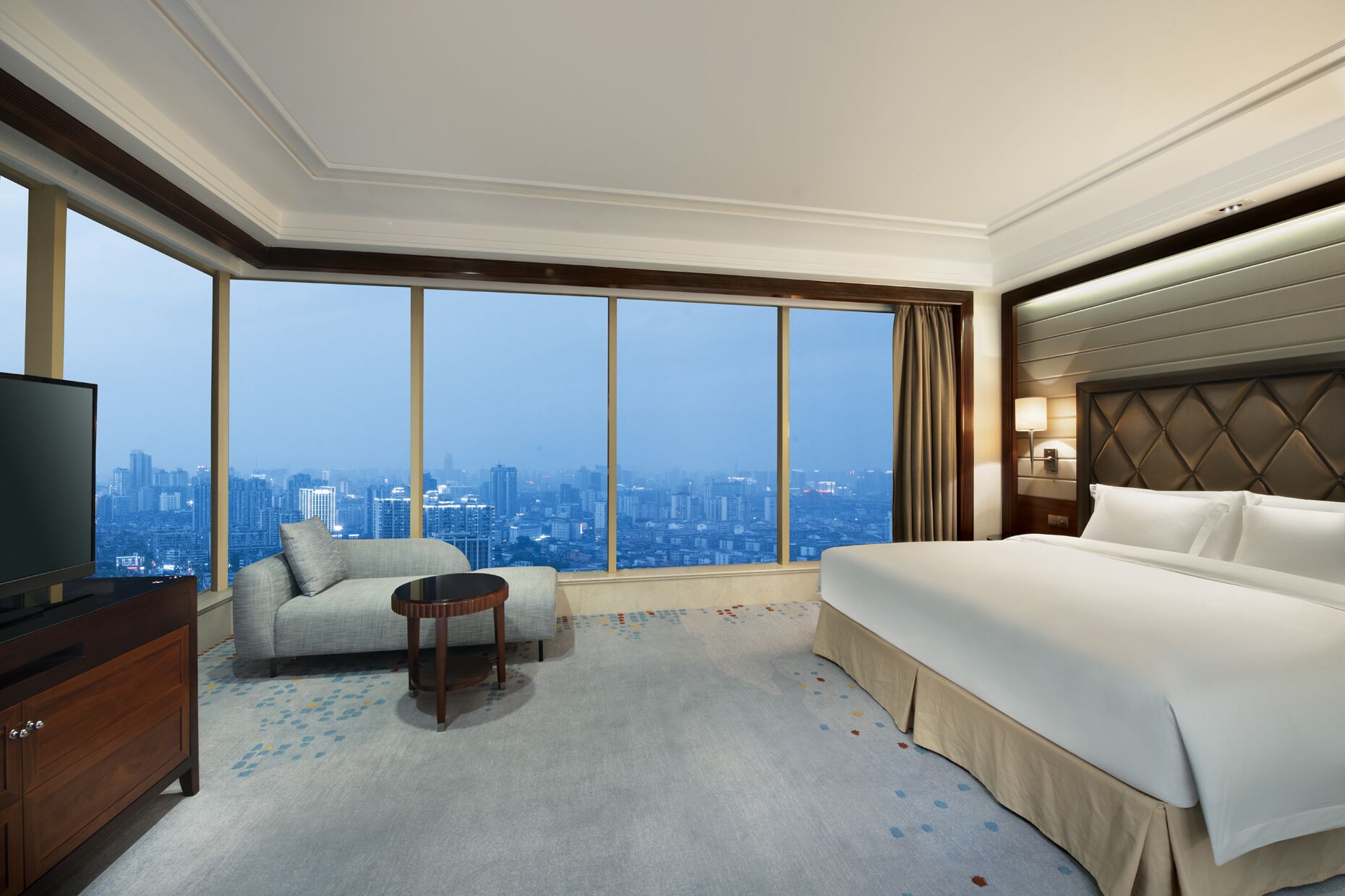 A hotel room with a large window, a bed, a sofa, a TV, and a small table.