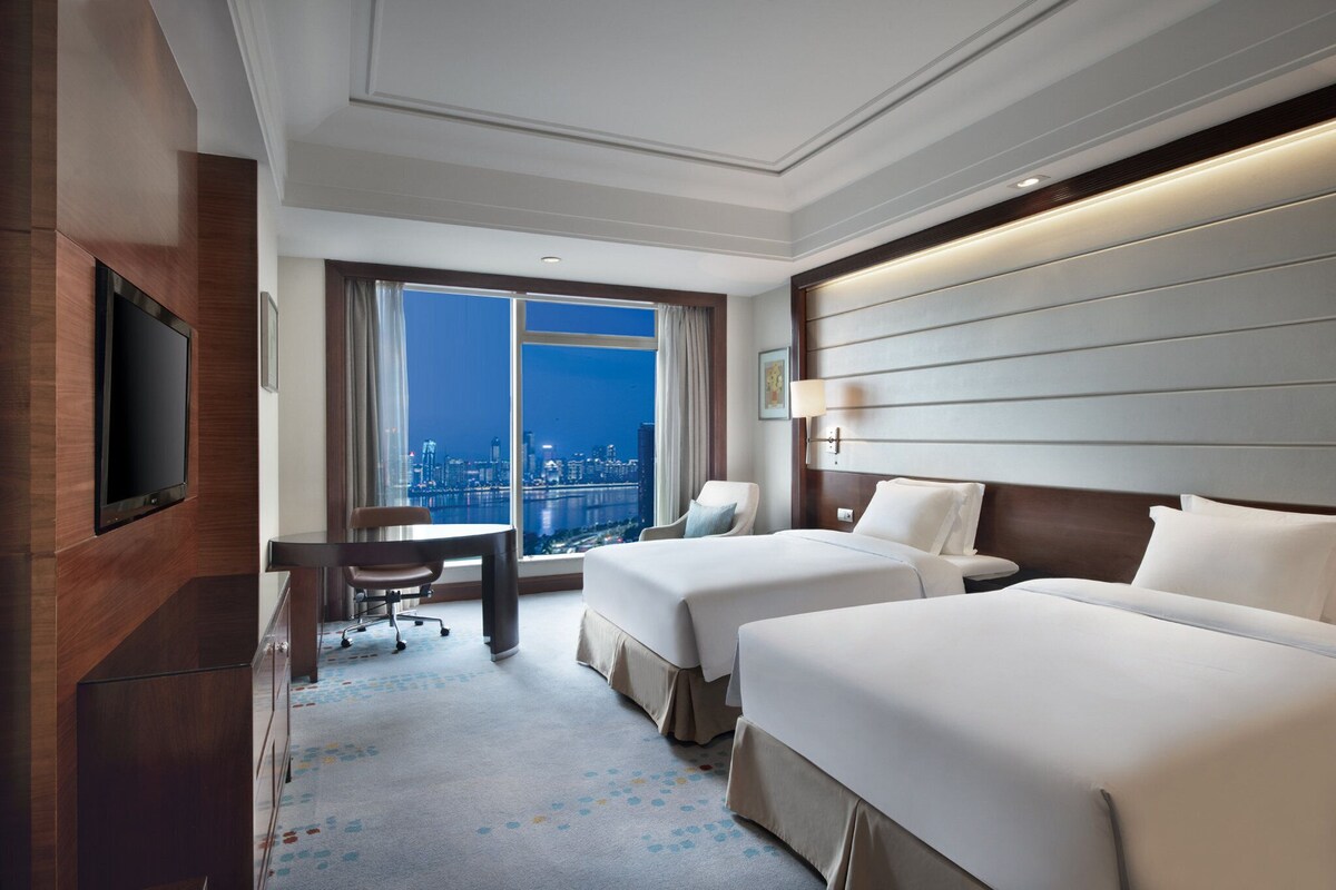 A hotel room with two beds, a desk, a chair, a TV, and a large window with a city view.
