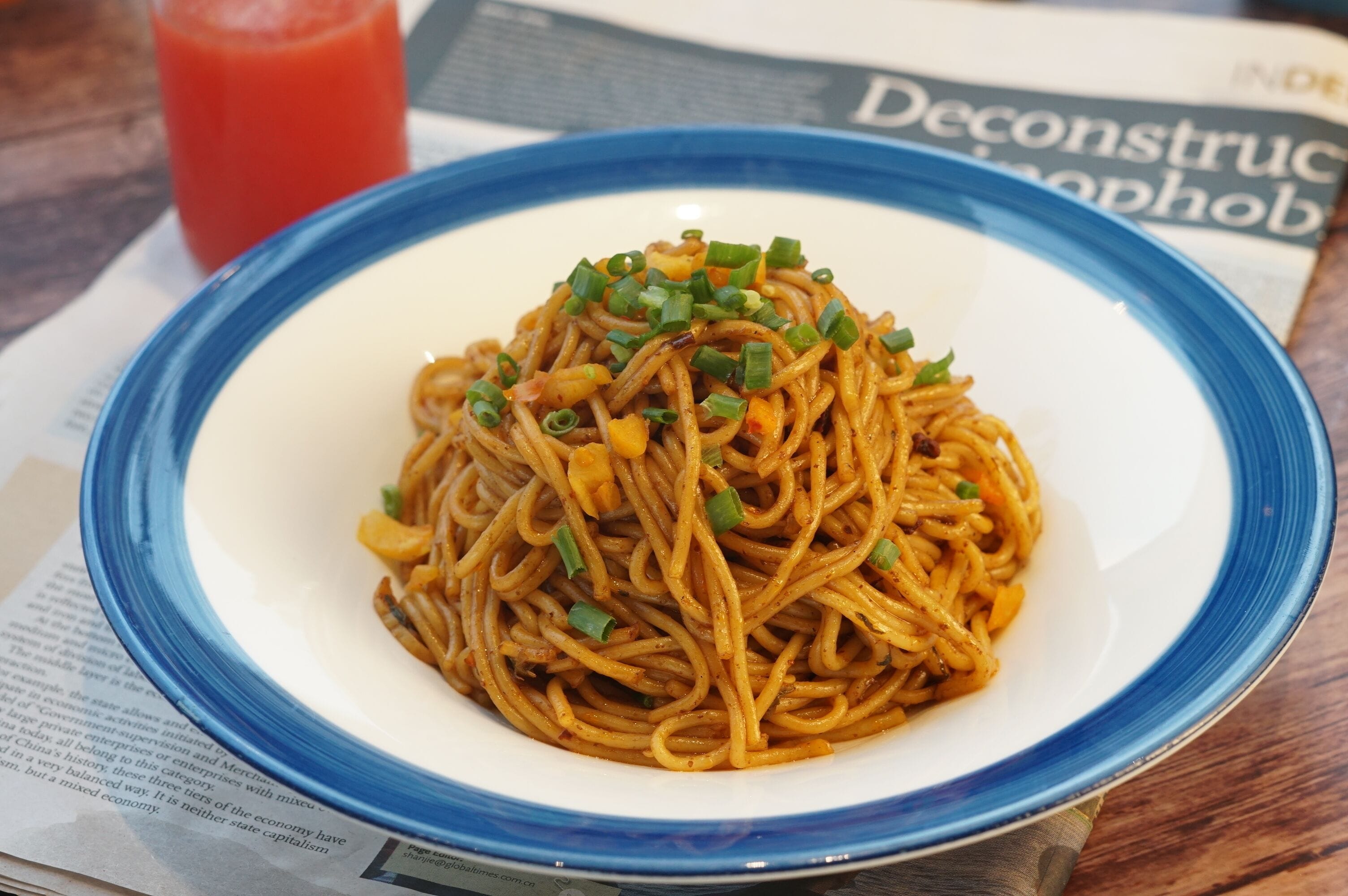 A plate of spaghetti with green onions on a blue-rimmed white plate, placed on a table with a newspaper and a glass of red drink.