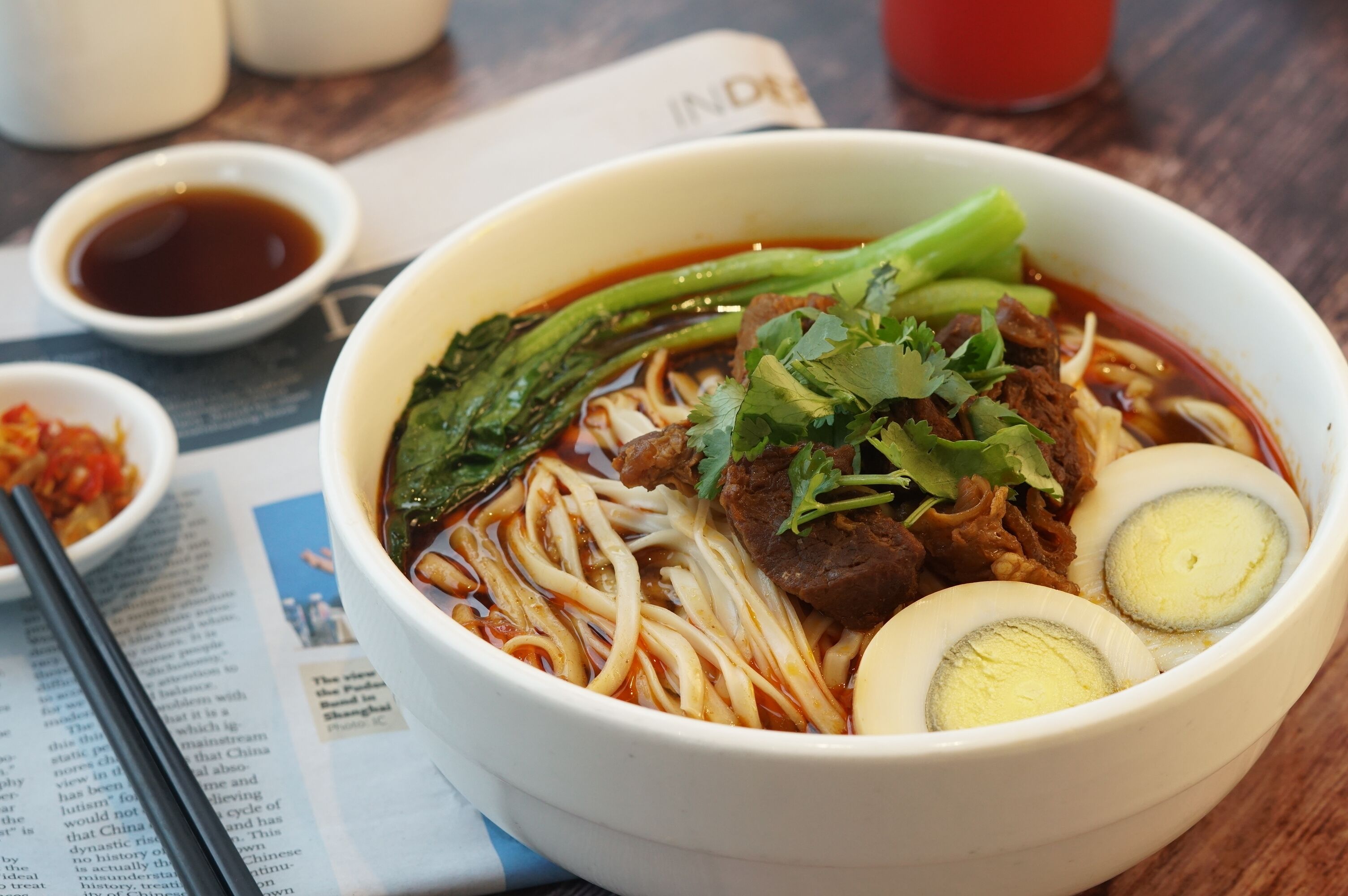 A bowl of noodle soup with beef, boiled eggs, and vegetables, accompanied by small bowls of sauces.