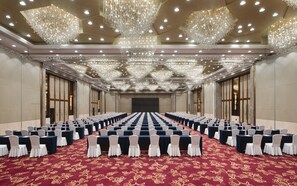 A large conference room with rows of tables and chairs, ornate chandeliers, and a red carpeted floor.