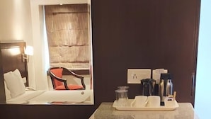 Une chambre d’hôtel compacte comprenant un petit coin cuisine, un lit avec une tête de lit, un espace salon avec un fauteuil et une fenêtre avec des stores.