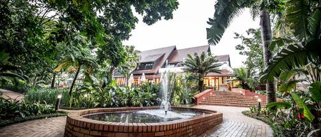 A courtyard with a brick fountain, lush greenery, and a building with a sloped roof in the background.