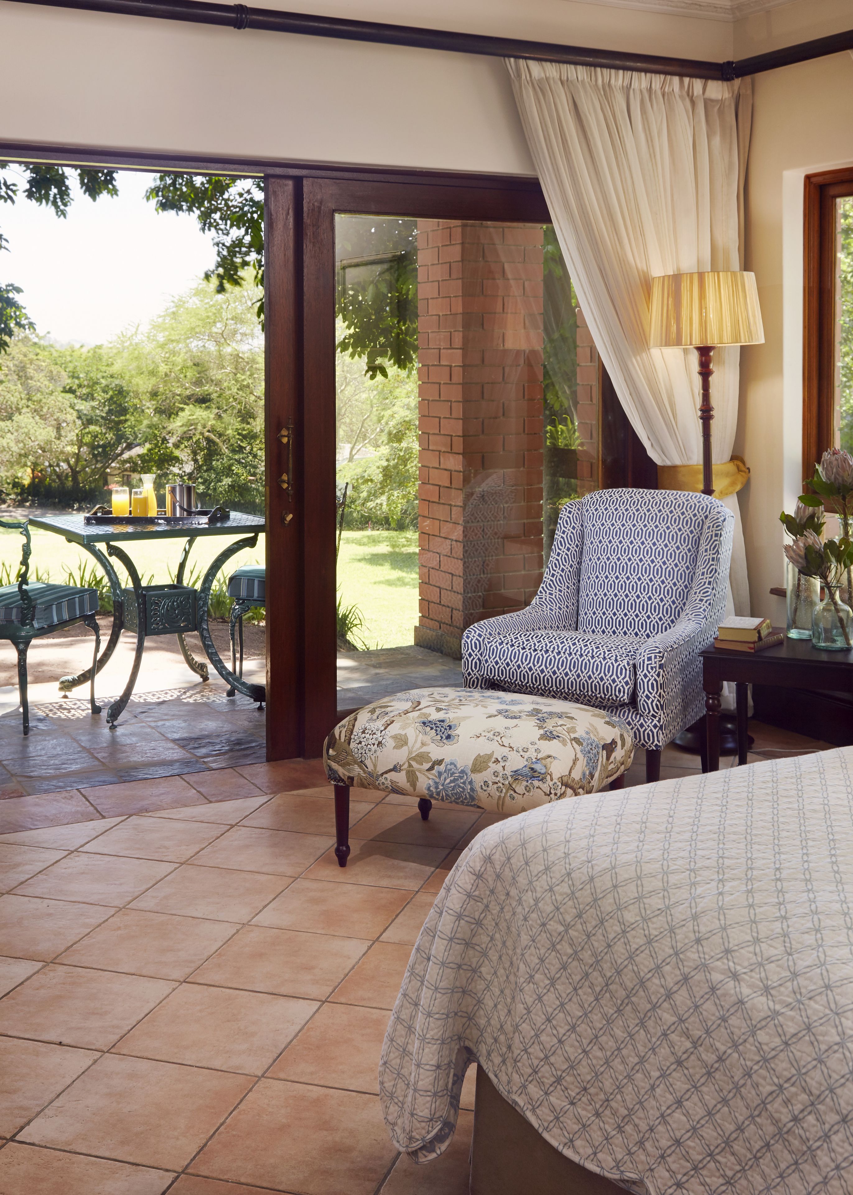 A bedroom with a bed, a patterned armchair, a table with a lamp, and a view of the outdoors.