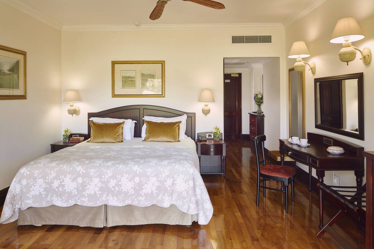 A hotel room with a large bed, two bedside tables, a desk with a chair, a mirror, and a ceiling fan.