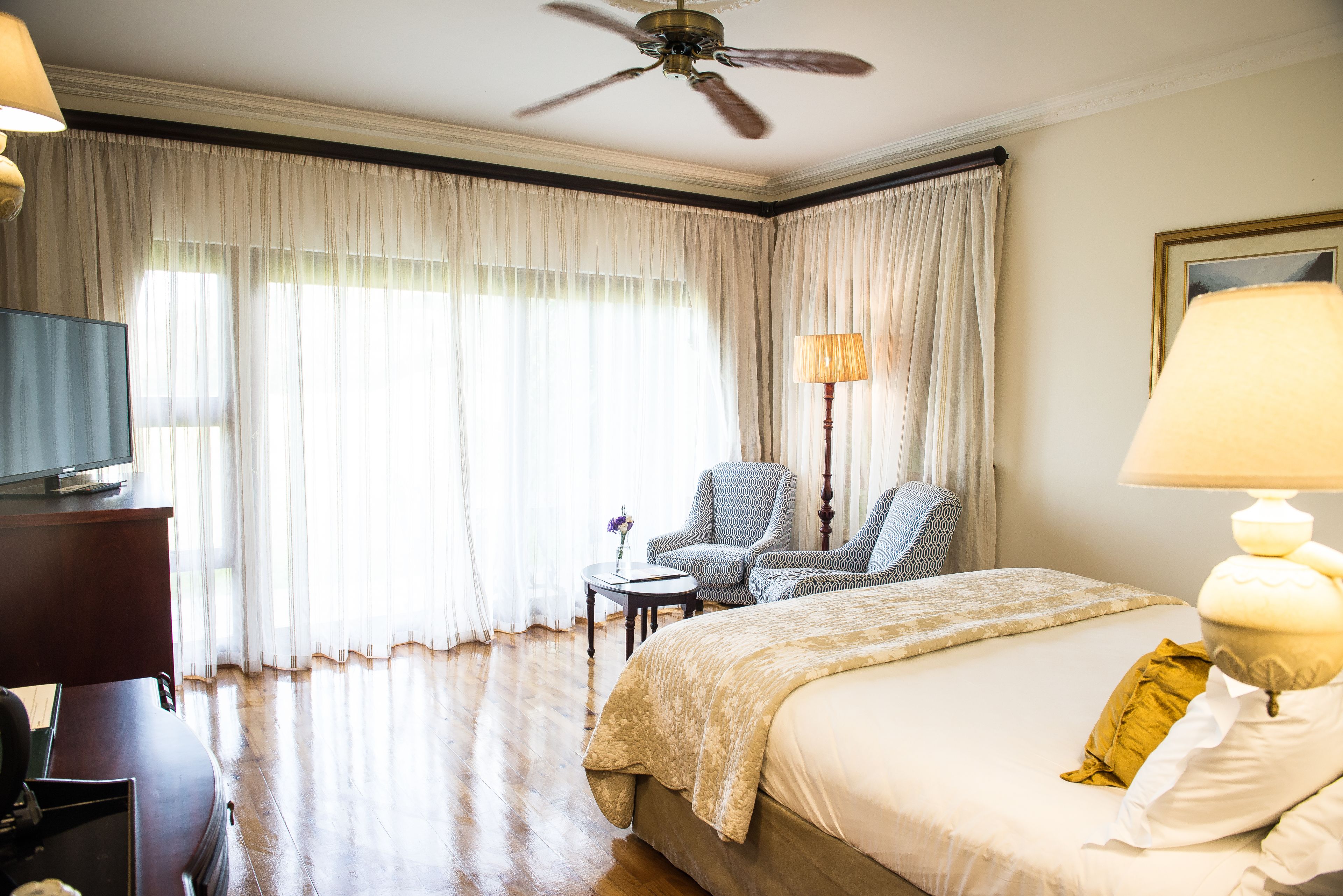 Ein Hotelzimmer mit Bett, Schreibtisch, Stuhl, Fernseher und Deckenventilator.