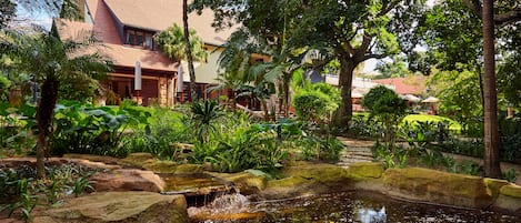 A tropical garden with a pond, lush greenery, and a building with a red roof.