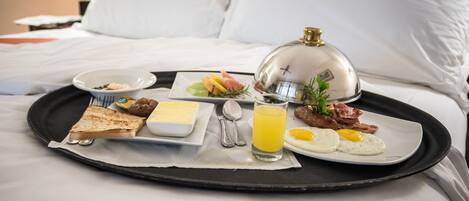 Bandeja de desayuno sobre una cama, con tostadas, huevos, tocino, un vaso de zumo de naranja y un plato pequeño de fruta.
