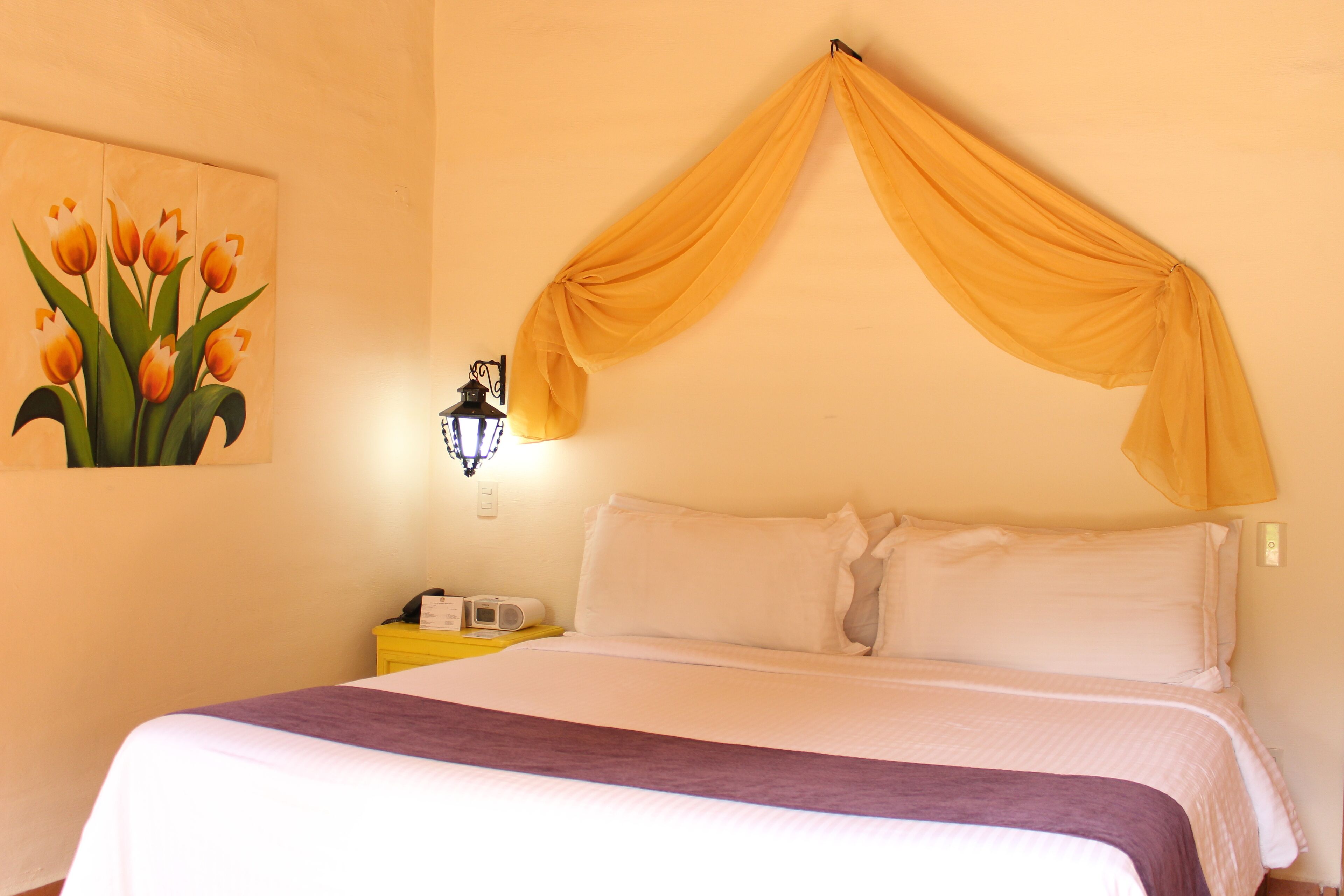 A bed with a canopy, a nightstand with a lamp, and a painting of tulips on the wall.