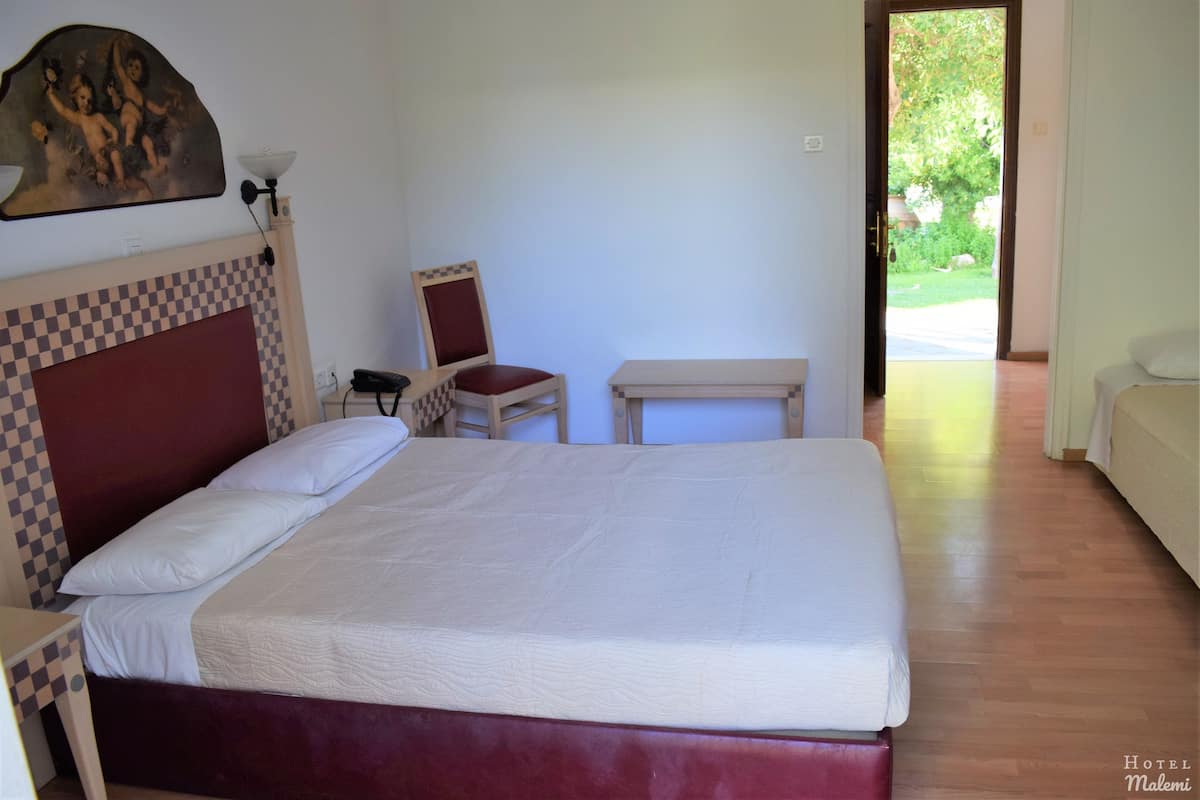 A hotel room with a checkered headboard, a bed with white linens, a small table, a chair, and a painting on the wall.