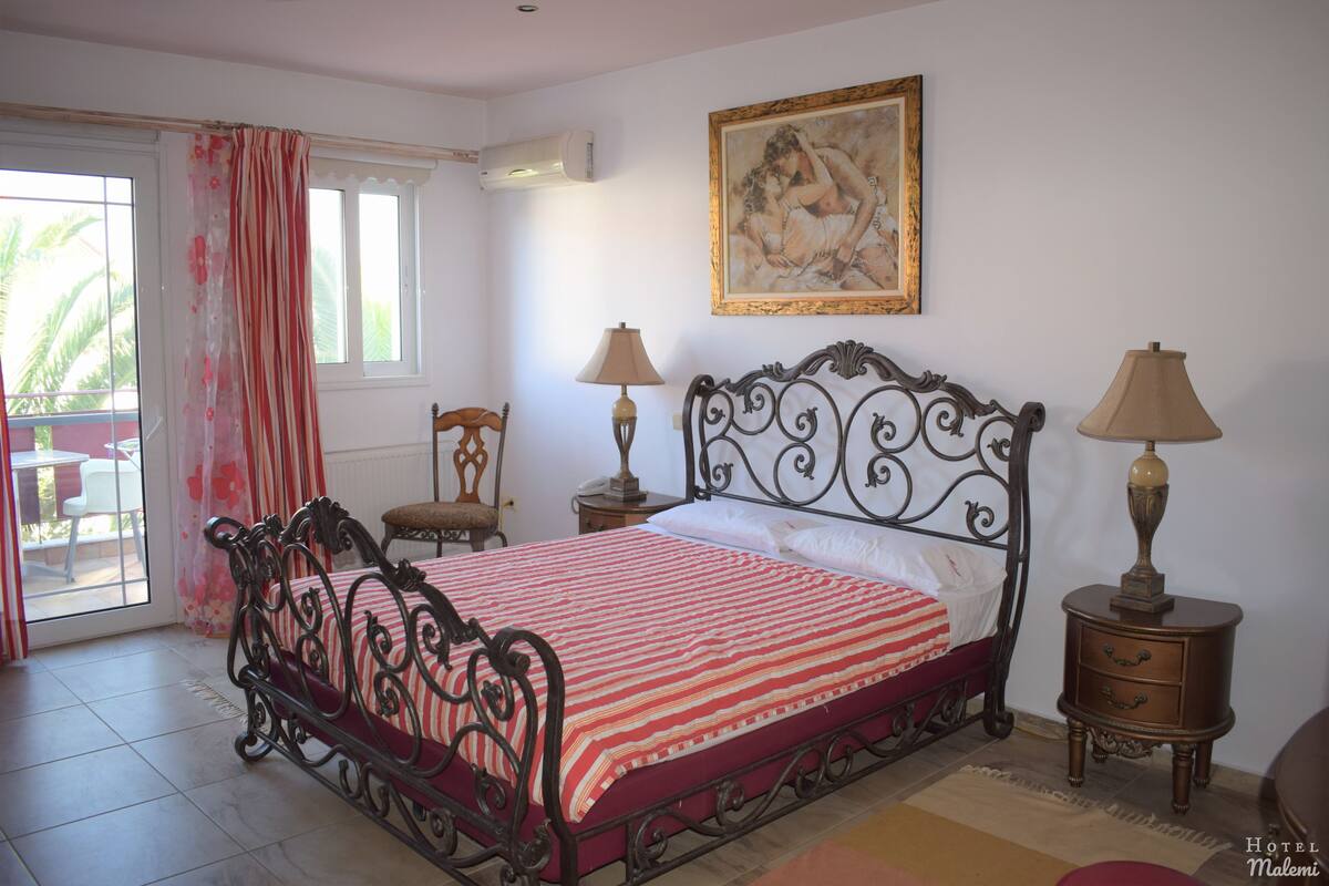 A bedroom with a metal bed frame, a red and white striped bedspread, two bedside tables with lamps, a chair, and a large painting on the wall.