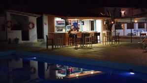 A bar area with outdoor seating, a pool in the foreground, and a building with a tiled roof. - Malemi Organic Hotel (Lesvos)