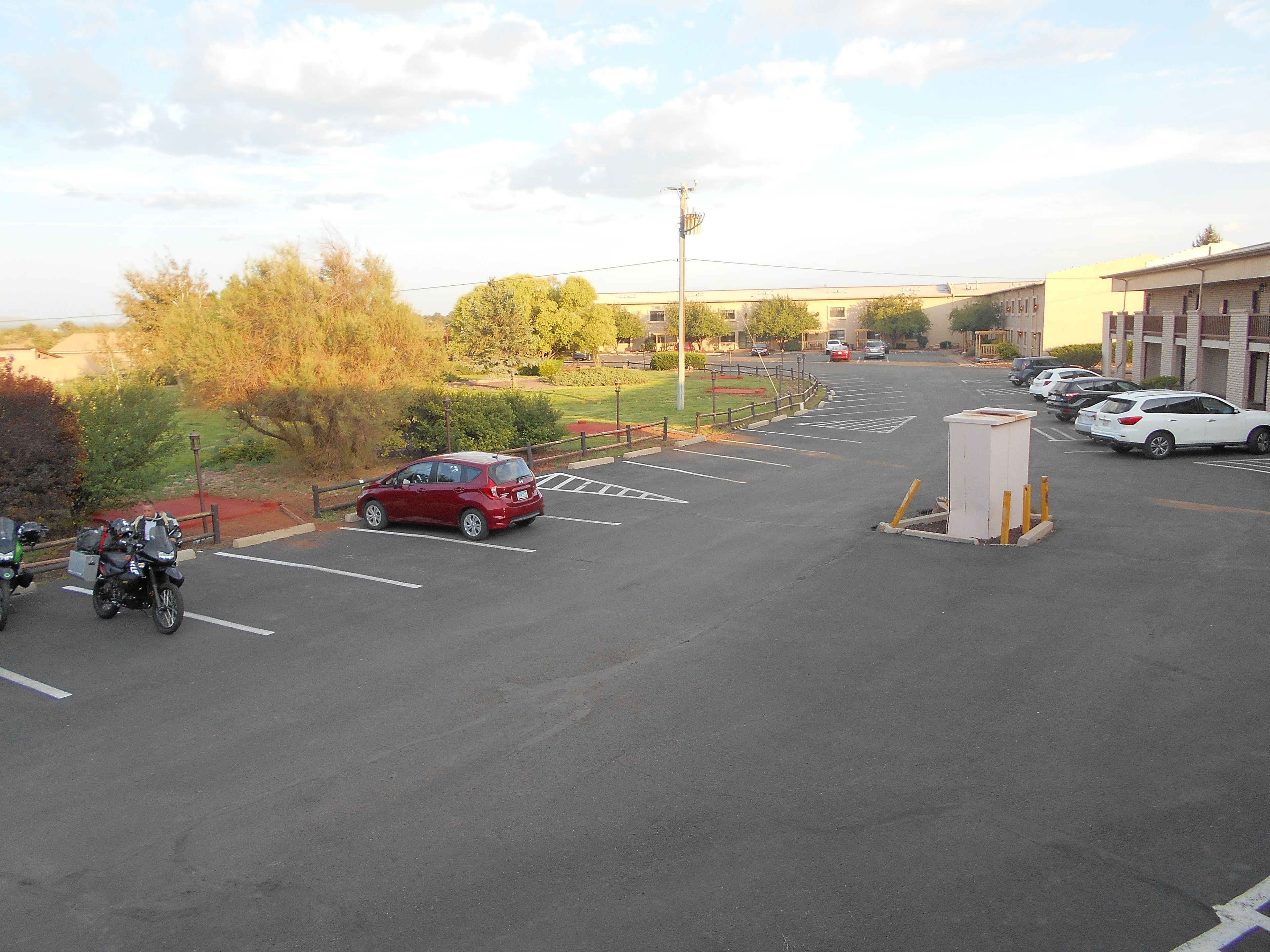 Een parkeerplaats met diverse auto's en motorfietsen, een gebouw op de achtergrond en een omheind gebied met bomen.