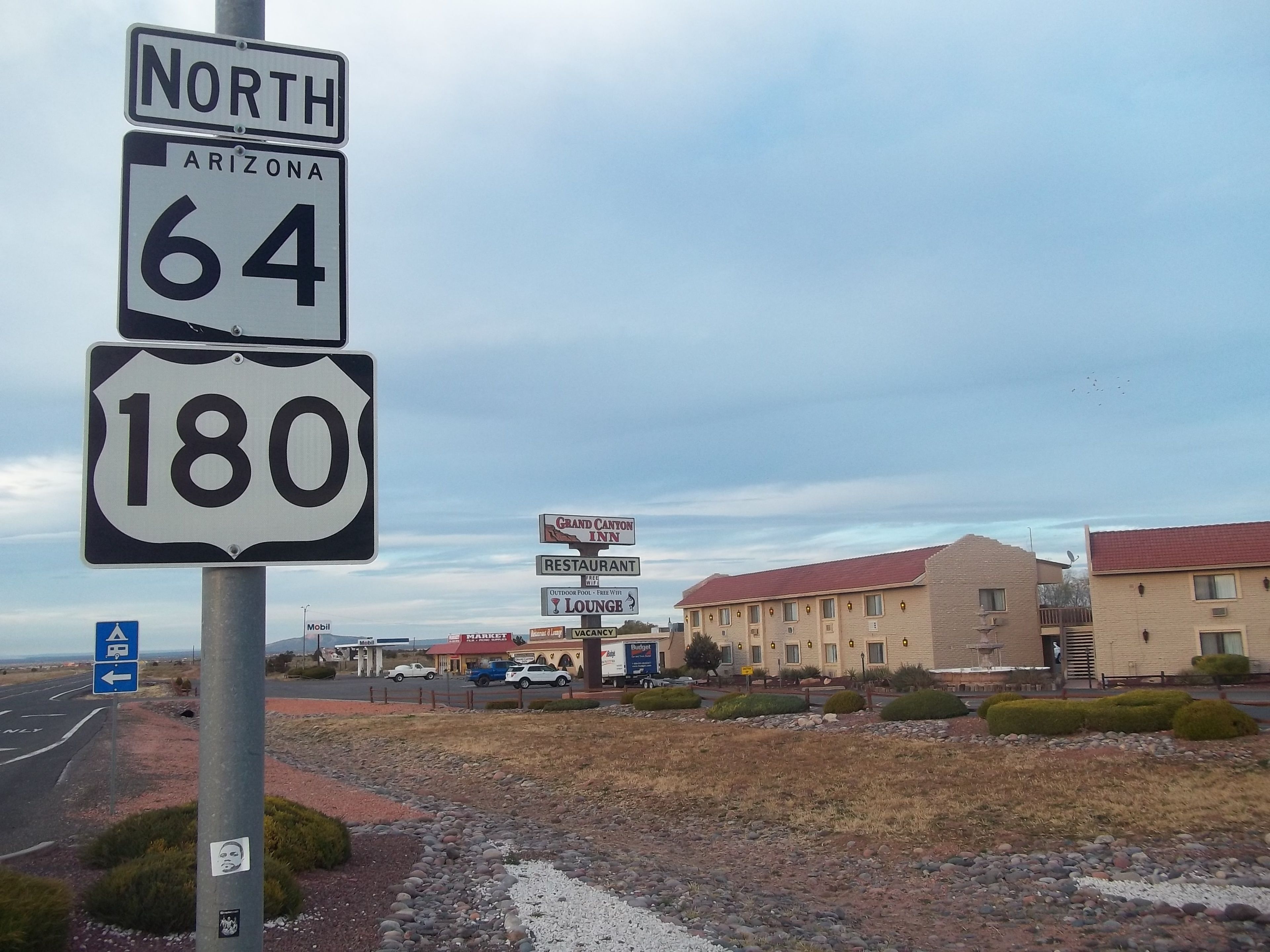 Un panneau de signalisation indiquant la direction de la route 64 Nord et de la route 180 en Arizona, avec un panneau d’un motel et d’un restaurant en arrière-plan.