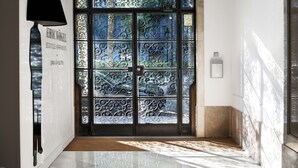 Un pasillo con una puerta negra que presenta detalles de metalurgia intrincada y paneles de vidrio, una lámpara fijada a la pared y un piso de baldosas.