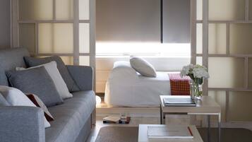 A modern hotel room with a grey sofa, a white bed, a small table with a book and a glass, and a window with blinds.