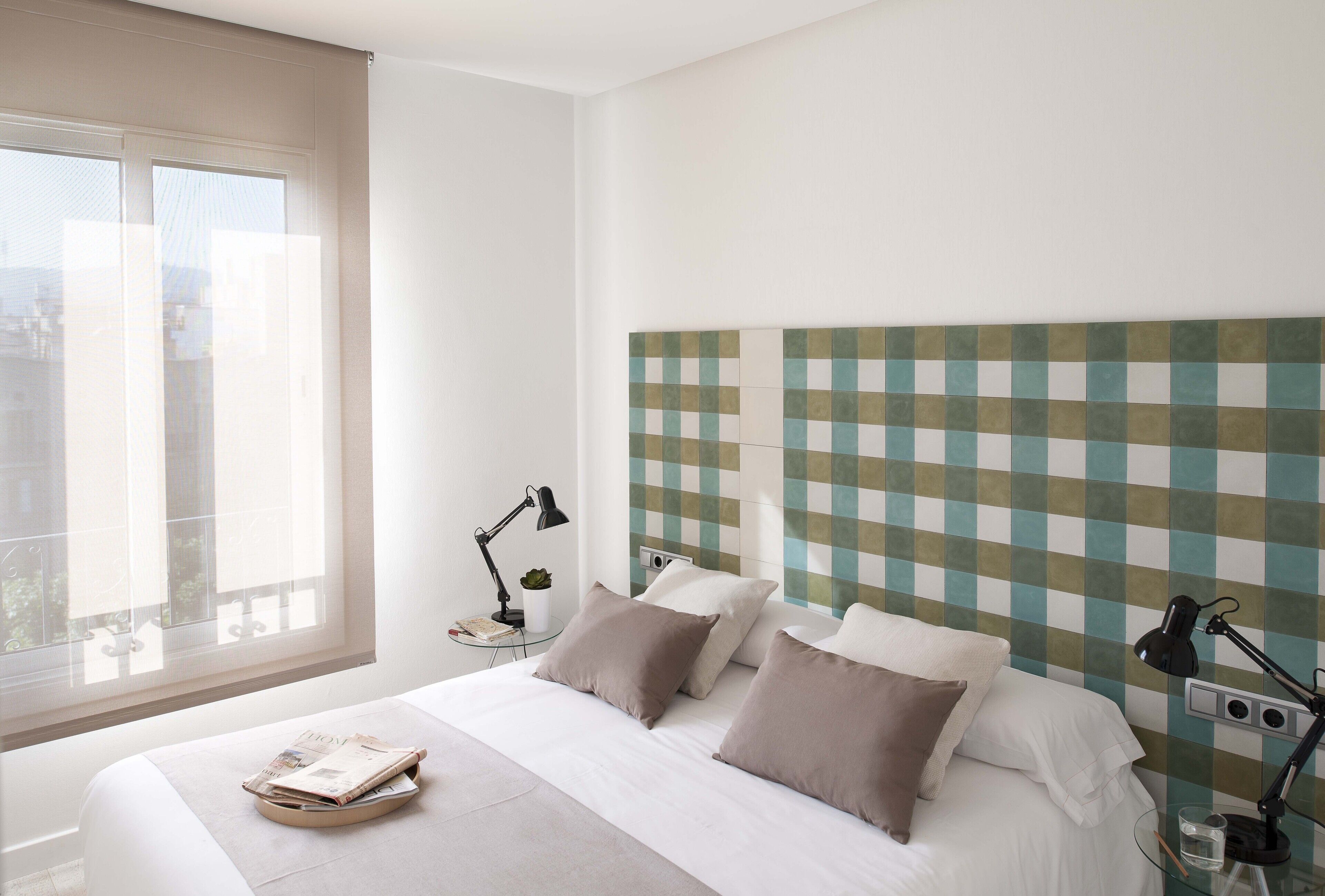 Una habitación de hotel moderna con una cama grande, cabecero a cuadros, mesita de noche con lámpara y ventana con vista a edificios.