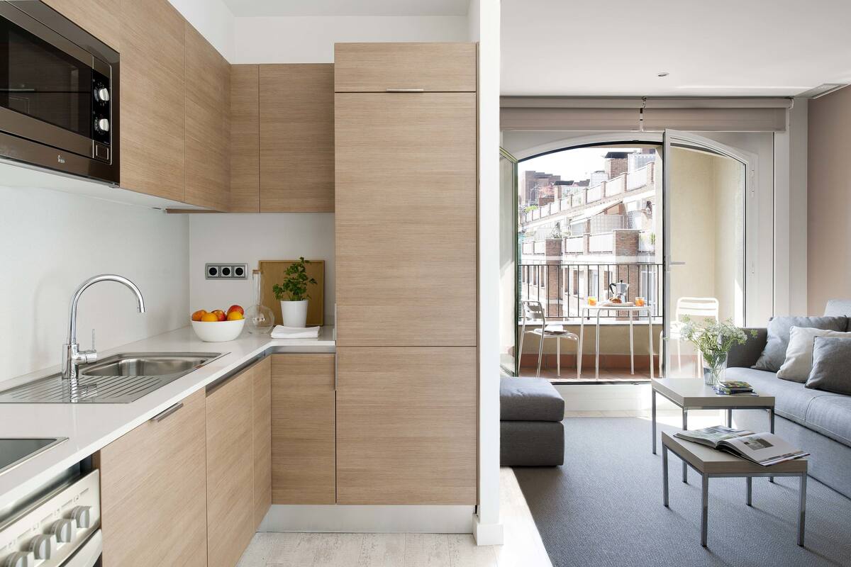 Cocina moderna con horno integrado, un lavamanos y un cuenco de fruta. Hay una mesa pequeña con una planta y un libro. La habitación tiene un ventanal con vistas a edificios.