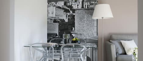 A modern living room with a grey sofa, a glass dining table with clear chairs, and a cityscape painting on the wall.