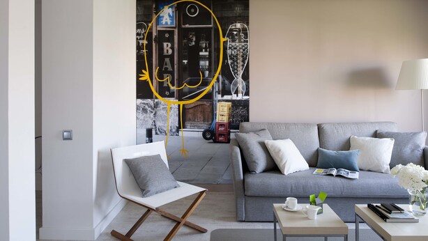 Una sala de estar moderna con un sofá gris, una silla blanca y una mesita de centro. Se observa una estantería con libros y objetos decorativos.