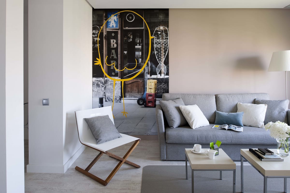 Una sala de estar moderna con un sofá gris, una silla blanca y una mesita de centro. Se observa una estantería con libros y objetos decorativos.