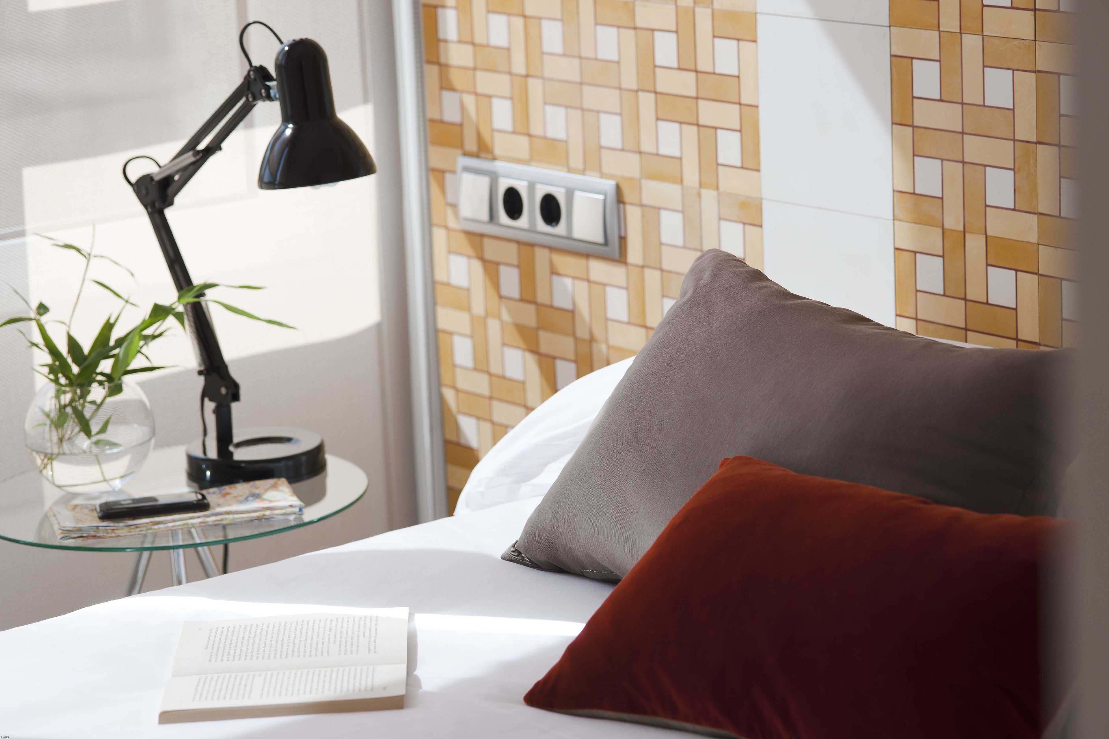 A neatly made bed with a white comforter and a red pillow, a glass table with a black desk lamp, a book, and a small plant.