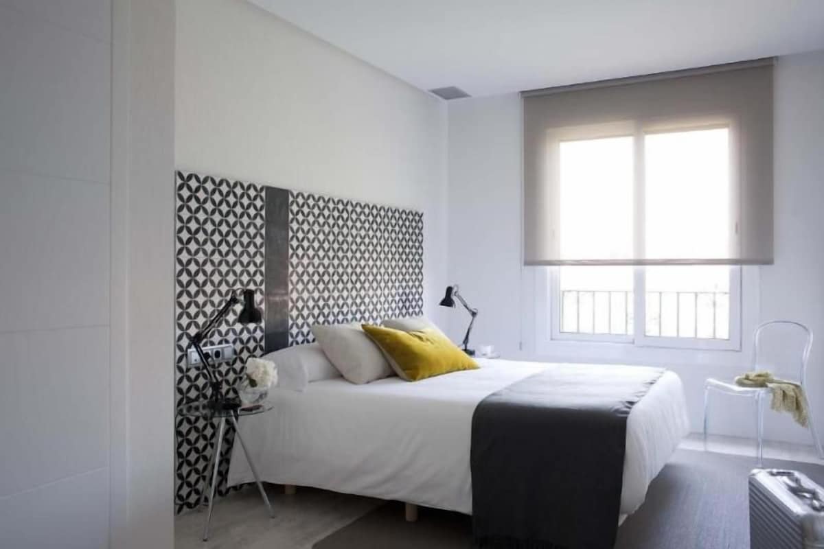 Un dormitorio moderno con un cabecero estampado, una cama con ropa de cama blanca y un almohadón amarillo, una mesita de noche con una lámpara y una ventana con persianas.