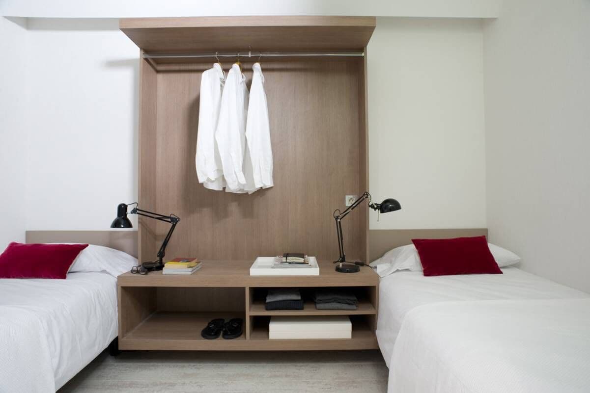 Un dormitorio con dos camas, un armario de madera, un escritorio con lámpara y un estante con zapatos.