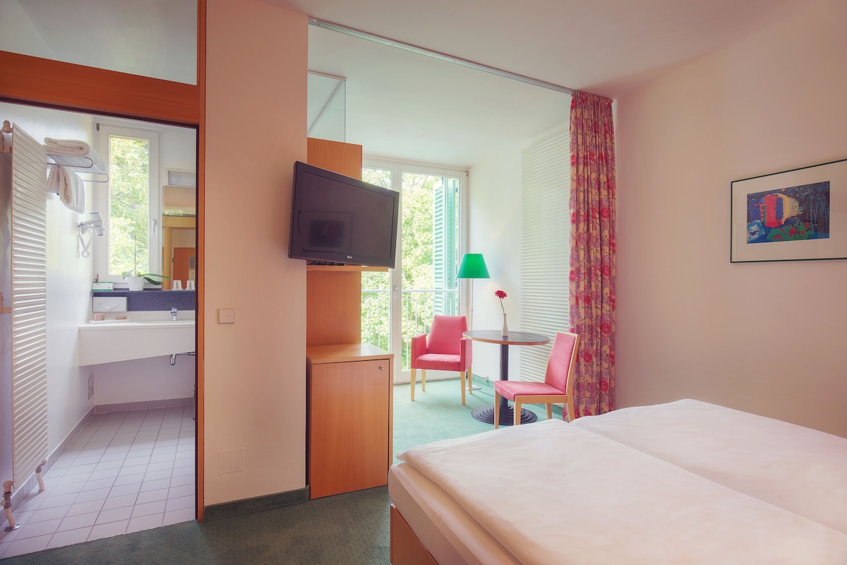 A hotel room with a bed, a TV, a small table with a chair, a bathroom with a sink and mirror, and a balcony with a chair and a table.