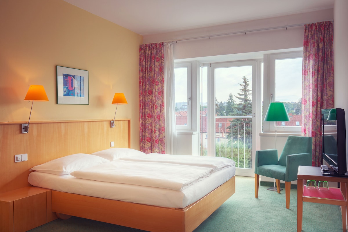 A hotel room with a bed, two lamps, a chair, a desk with a computer, and a window with curtains.