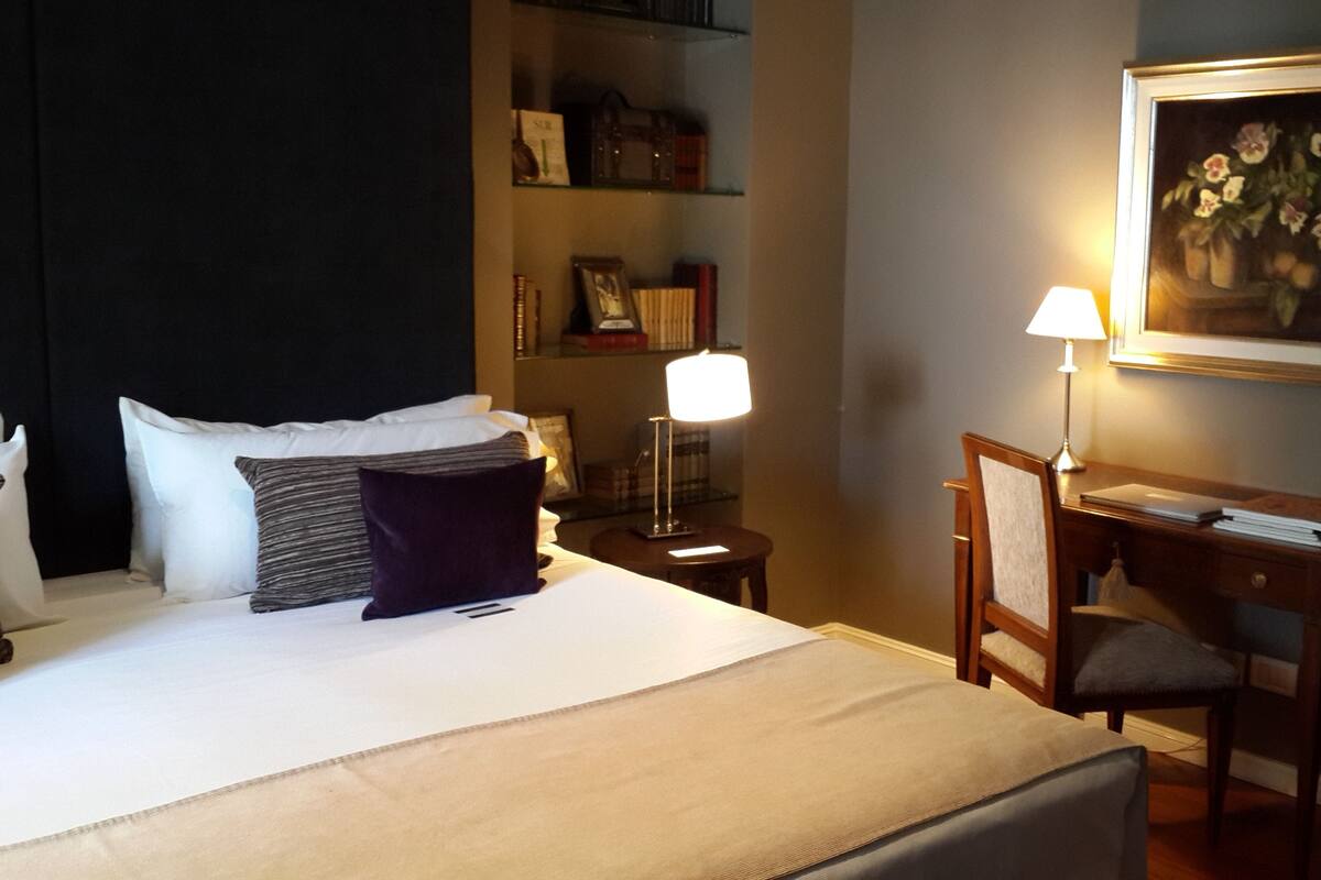 A hotel room with a bed, a desk, a chair, a lamp, a bookshelf, and a framed painting on the wall.