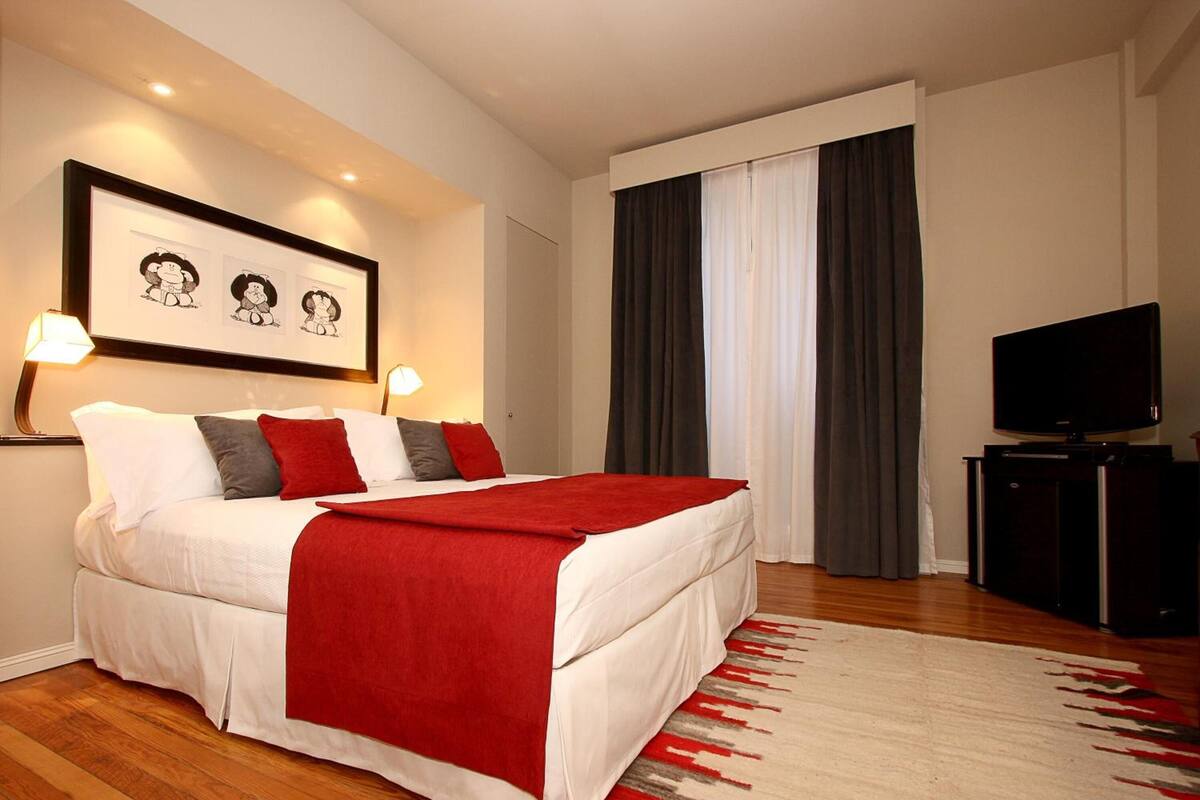 A neatly made bed with a red blanket, a framed picture above it, and a television on a stand to the right.