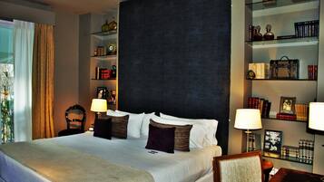 A hotel room with a large bed, two bedside tables with lamps, a dark headboard, and shelves with books and decorative items.