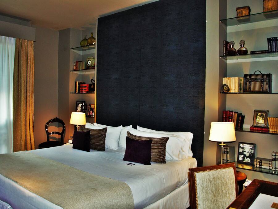 A hotel room with a large bed, two bedside tables with lamps, a dark headboard, and shelves with books and decorative items.