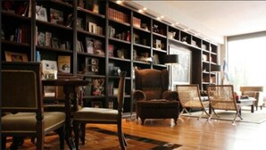Una sala de estar acogedora con piso de madera, una amplia estantería llena de libros, un sillón de cuero y una silla de mimbre.