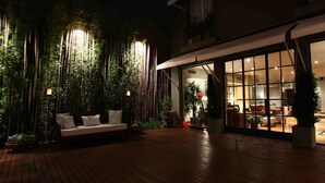 Terraza de madera al aire libre con un sofá blanco y plantas en macetas, que conduce a una puerta de cristal que da paso a un espacio interior bien iluminado.
